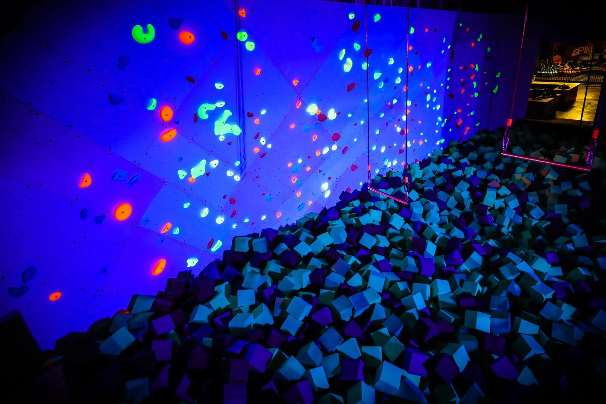 A view of the bouldering wall + foam pit finish at dark under blacklight. Even the foam looks great under blacklight, and there was enough room left over in the ex-swimming pool to accommodate kids climbing or swinging from trapeze bars.