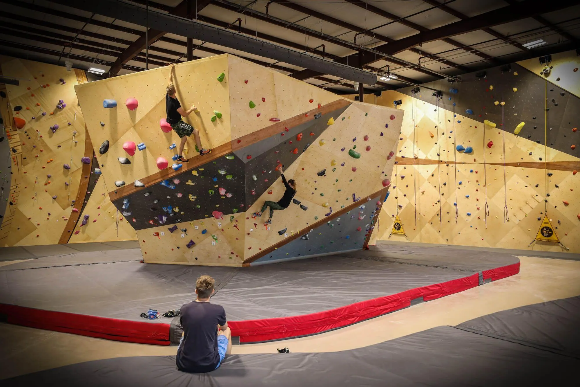 Two climbers solving problems while one more looks-on at the main boulder feature here at this indoor climbing gym. Unified by design with the route climbing walls, this boulder is one of the most popular features in the gym due to its vast variety in natural wall angles and problems that results. Also of note is the super-clean Habit Flooring install for both bouldering and route climbing.