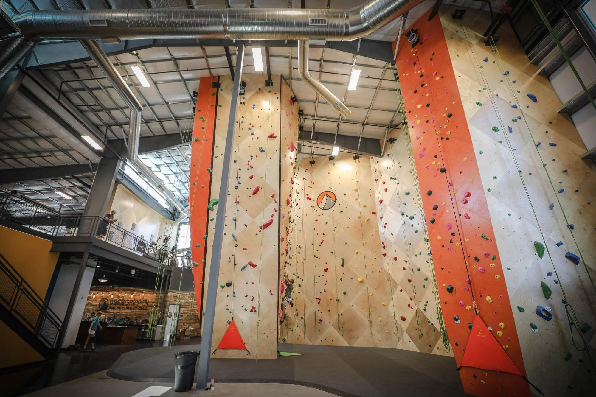 Standing in the middle of the gym, you can see the relationship between the entry/front desk, where it opens up into the route wall area, as well as one of the main bouldering areas above. Also prevalent is the gym logo front and center!